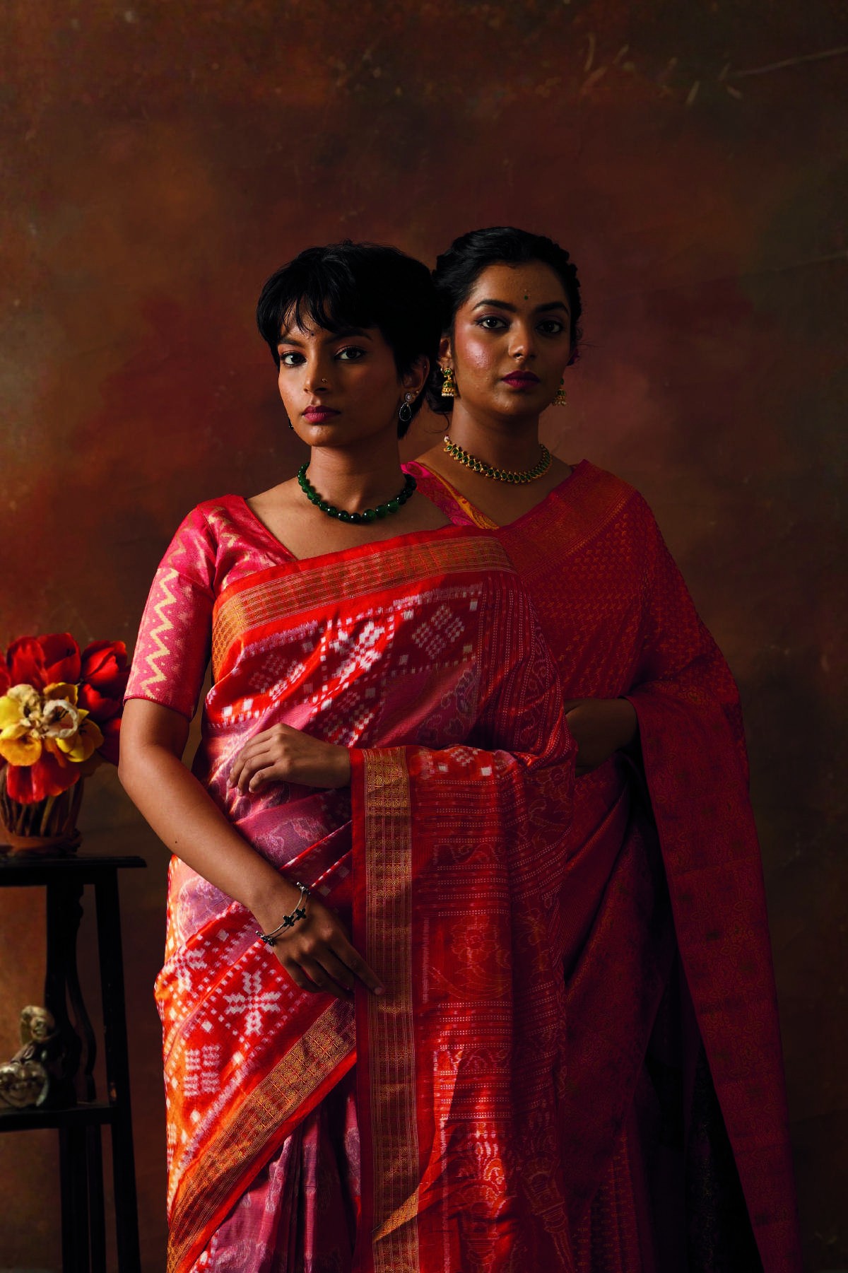 Two women in red silk sarees in a rich studio setting